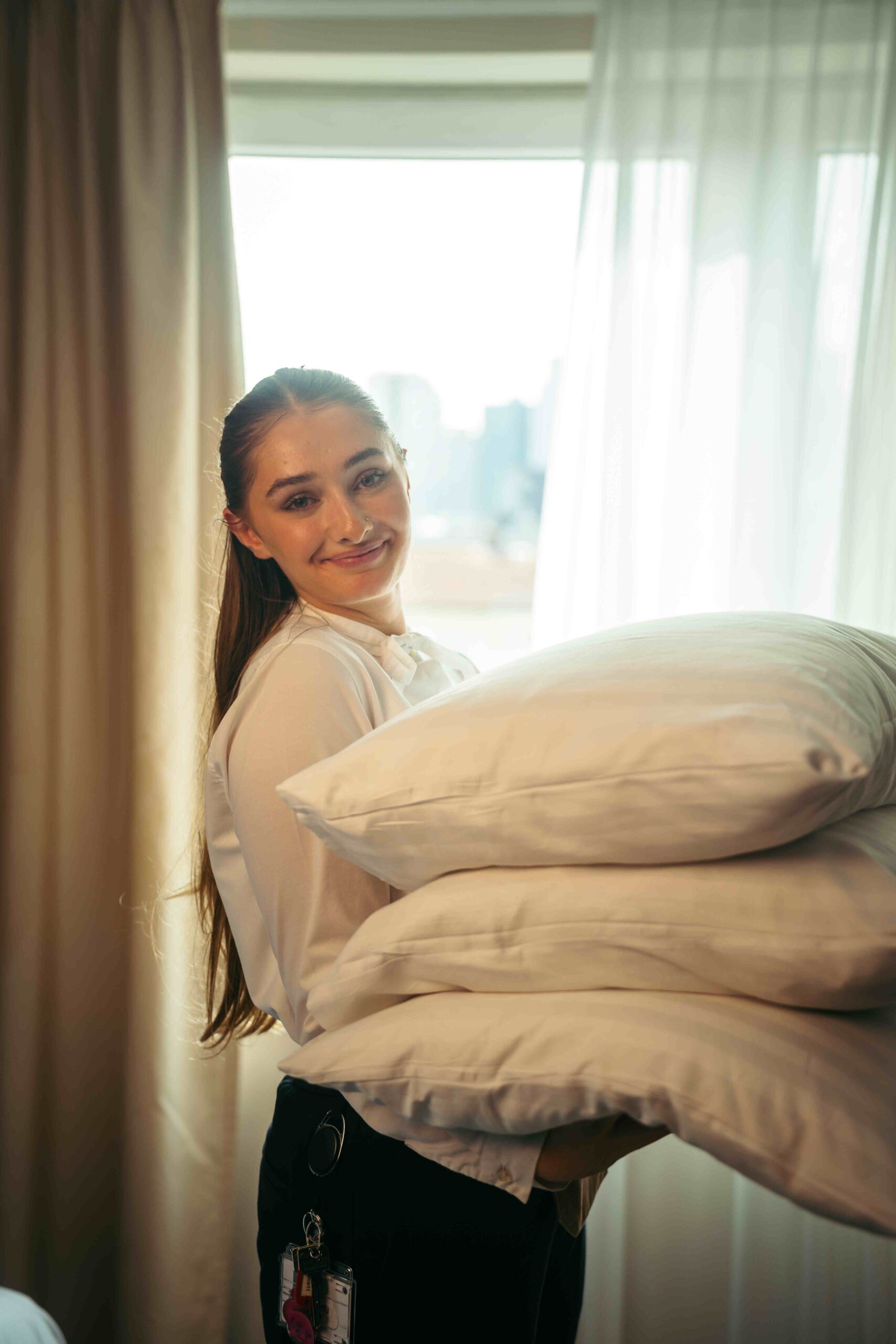 A smiling roommaid holding pillows infront of a window